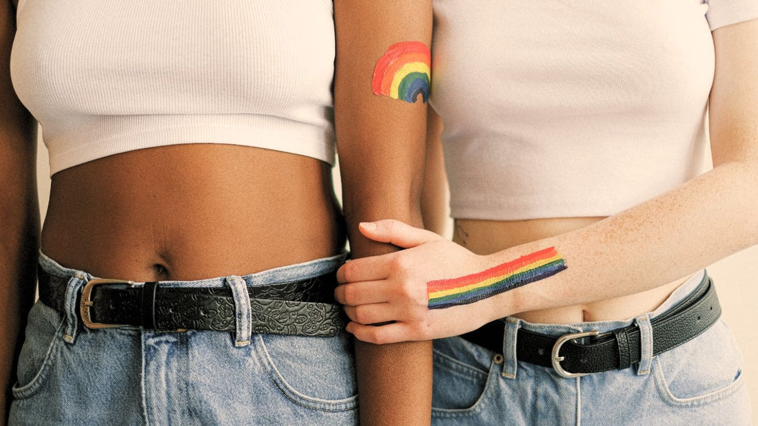 Deux femmes en jeans avec un haut blanc et le ventre découvert. Celle de droite tient le bras de celle de gauche. Elles ont des arcs-en-ciel LGBTQ+ dessinés sur le bras